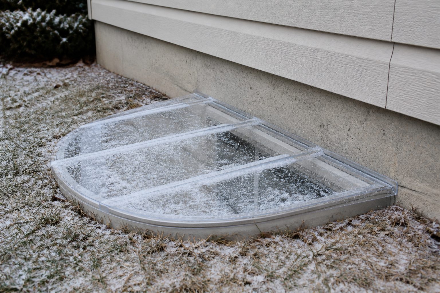 Clear window well cover with a light dusting of snow on a suburban foundation.